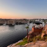 Wide-angle photo of the Nile River in Aswan, showing Elephantine Island with traditional Nubian homes and palm trees, framed by clear blue water and sky.