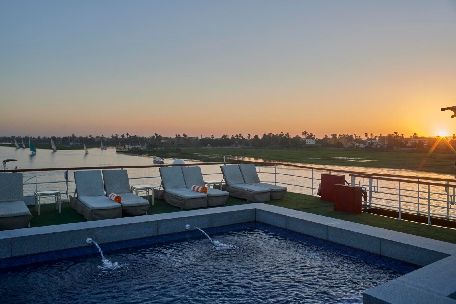 The sundeck and swimming pool of the luxurious Movenpick Royal Lily cruise ship, with a panoramic view of the Nile River's green banks under a clear blue sky.