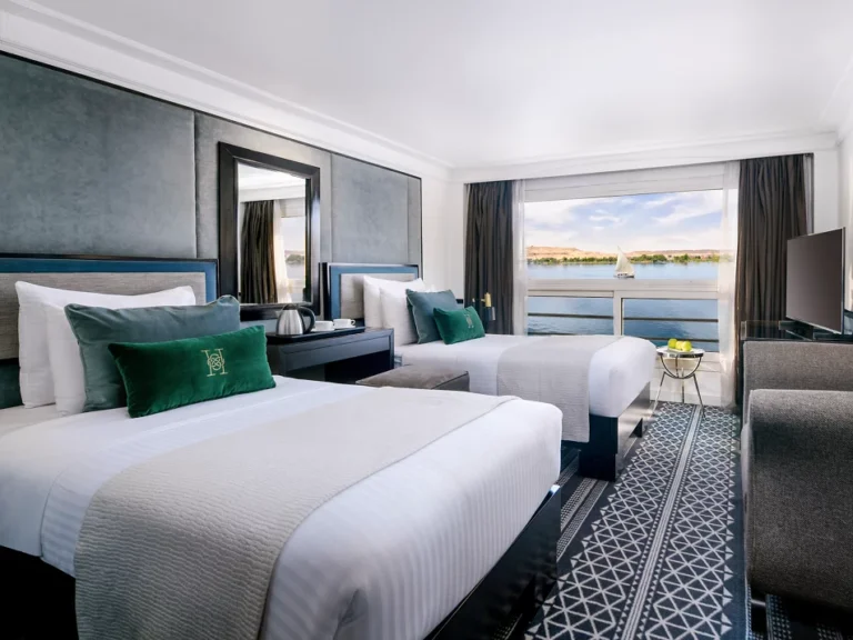 An opulent and spacious luxury cabin on the Movenpick Hamees Nile Cruise, featuring elegant furnishings, a comfortable bed, and large windows offering panoramic views of the Nile River