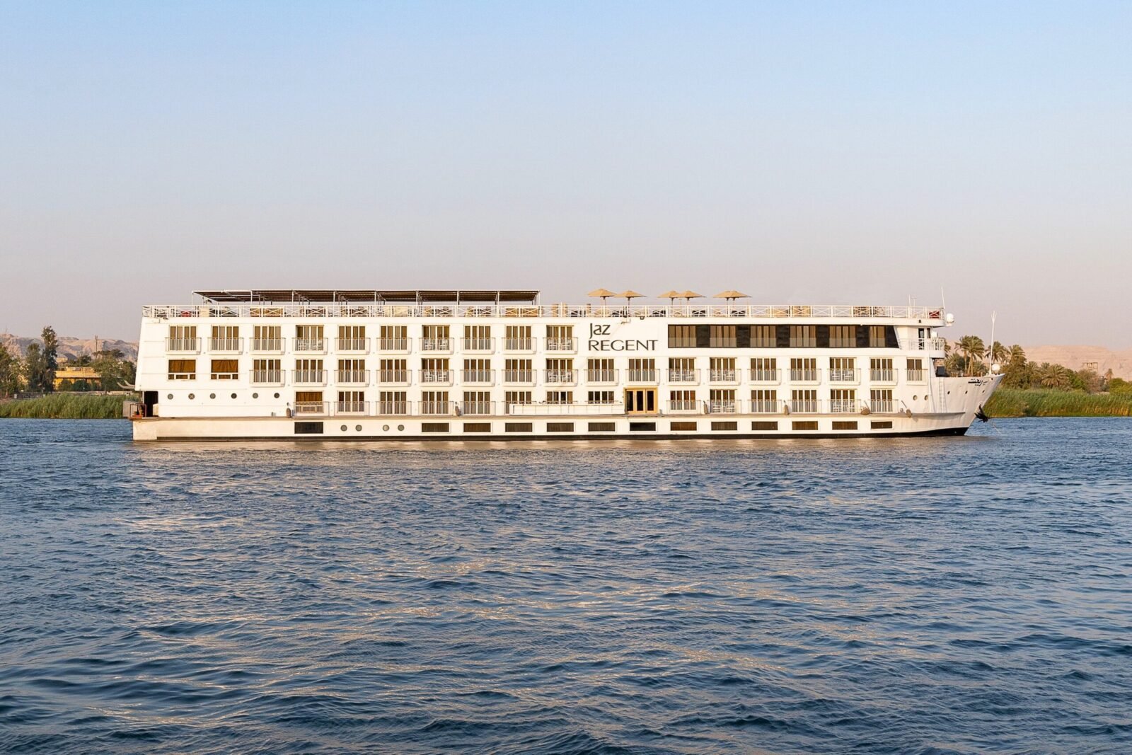 Photo of the large, modern Jaz Regent Nile Cruise ship sailing on the wide, blue Nile River with green banks visible in the distance