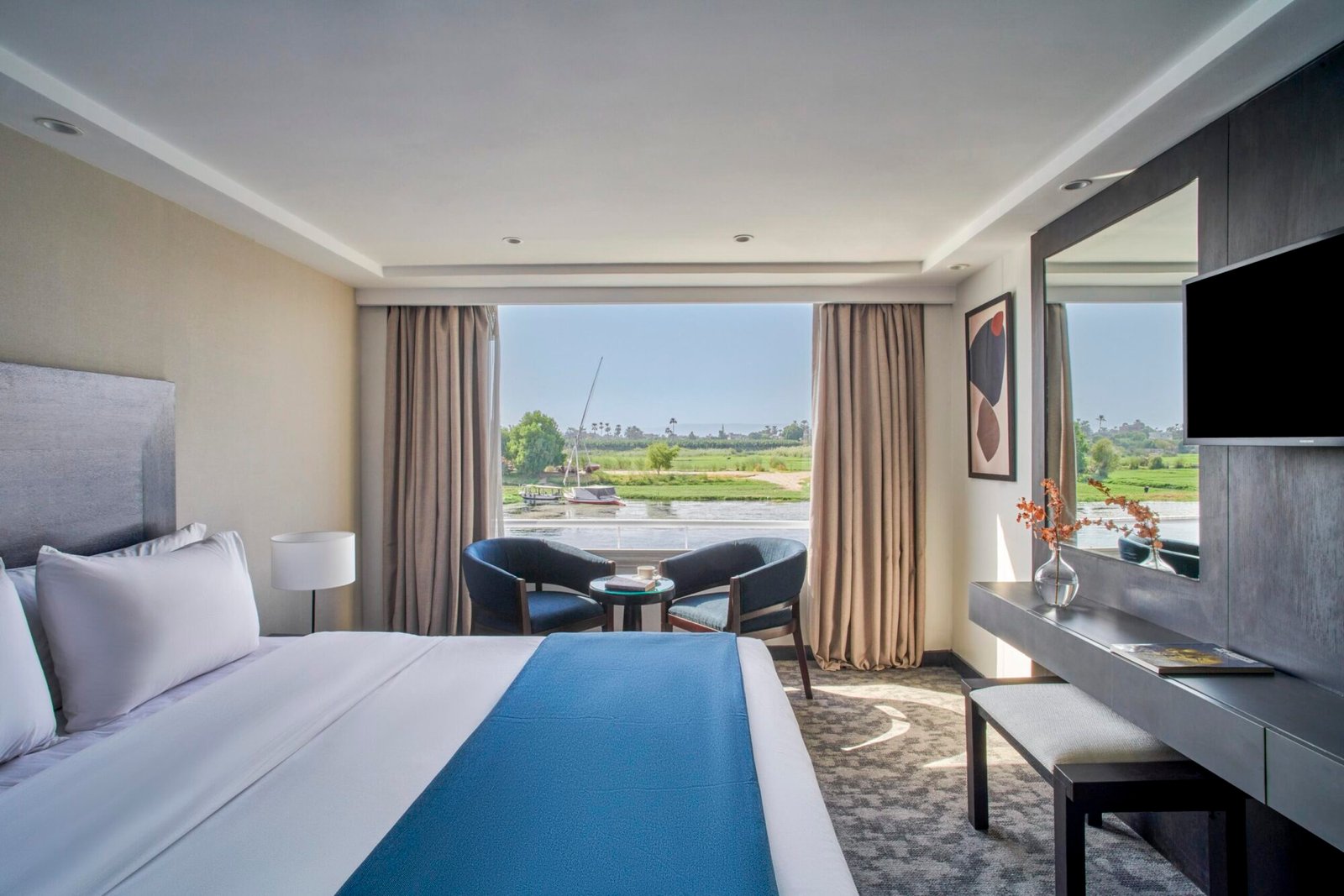 The interior of a beautifully decorated, luxurious cabin on the Movenpick Royal Lily Nile cruise, with a large, comfortable bed and a window looking out onto the Nile River.