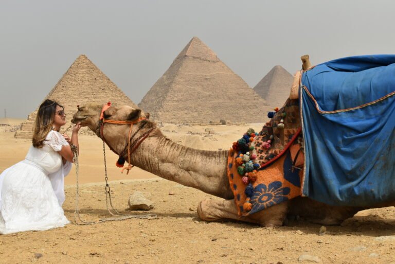 A female tourist enjoying a traditional camel ride with the Great Pyramids of Giza in the background under a clear blue sky.