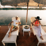 A couple relaxing on a sun lounger by the pool on a luxury Nile cruise sundeck at sunset, with the tranquil river and green banks in the background.