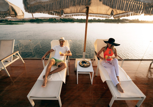 A couple relaxing on a sun lounger by the pool on a luxury Nile cruise sundeck at sunset, with the tranquil river and green banks in the background.