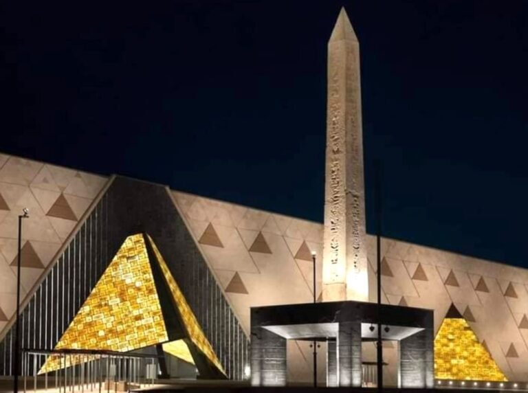 The Grand Egyptian Museum entrance at dusk, featuring the illuminated, modern building facade, the monumental Suspended Obelisk of Ramses II, and a clear call to action to book an online ticket or a combined Nile Cruise package.