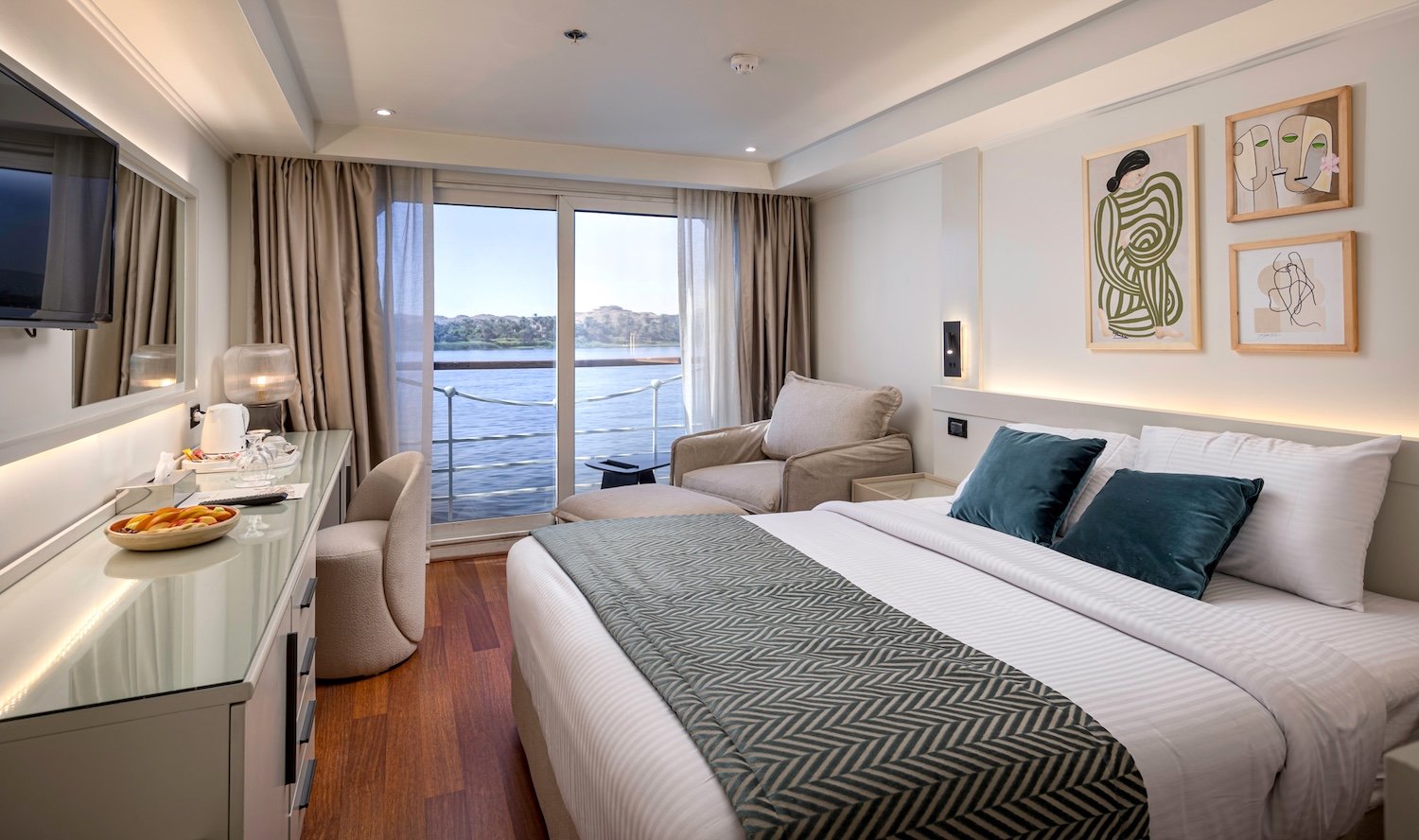 A modern, luxurious Grand Mandarin Nile cruise cabin showing a large panoramic window and balcony with a view of the Nile River and green banks outside.
