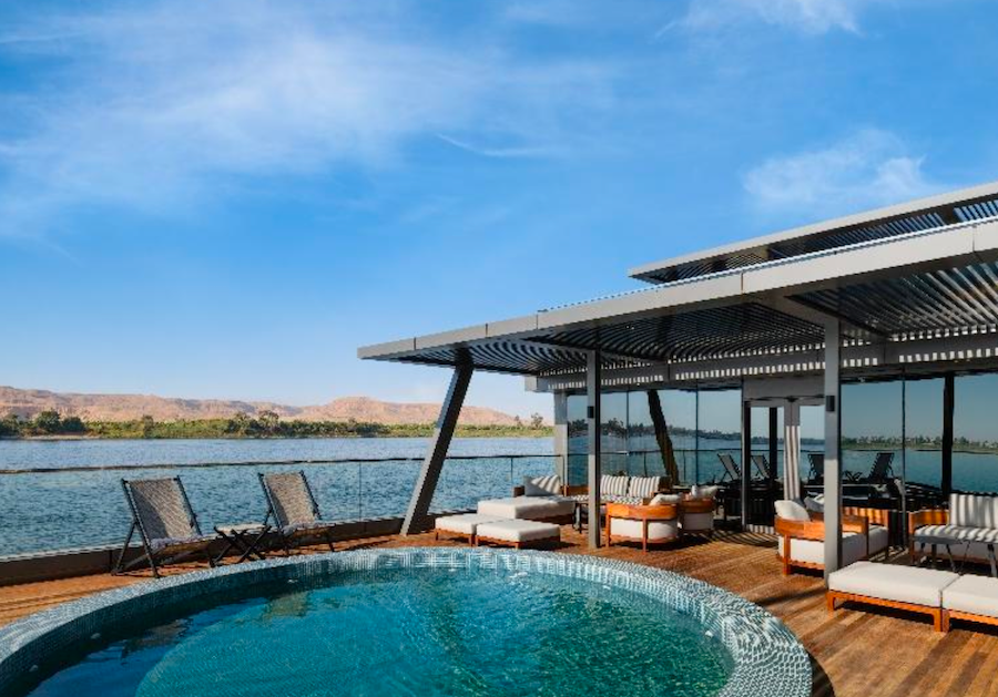 The wooden sun deck of the luxury Storia Dahabiya featuring a refreshing open-air plunge pool, white sun loungers, and a shaded canopy overlooking the Nile River